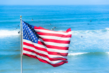 America flag blowing in the wind on blue Pacific ocean background with surfers silhouettes in...