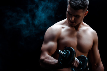 Fototapeta na wymiar sexy muscular bodybuilder with bare torso excising with dumbbell on black with smoke