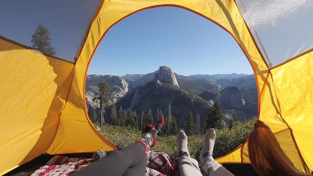 Camping rest in Yosemite Valley. A couple lay inside the yellow tent and point their fingers at the famous Half Dome rock that is seen outside on a picturesque mountain ridge. Yosemite National Park