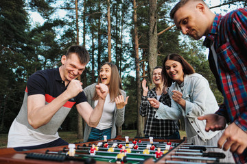 Friends play table football or kicker outdoors. Players and fans rejoice in the victory. - obrazy, fototapety, plakaty