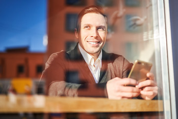 Happy male looking at camera with phone in hand sitting in cafe, reflection of building in glass