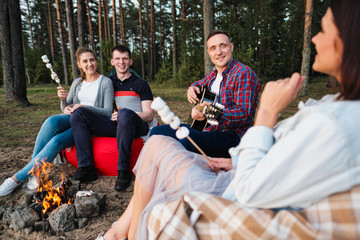 A group of friends relax in a forest camp. Men and women prepare a marshmallow on a bonfire. A party in nature.
