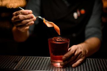 Bartender adding to the alcoholic drink spiral orange zest with tweezers