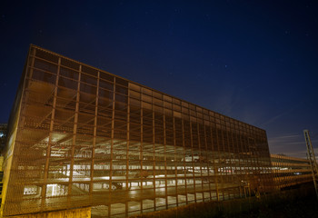 Fototapeta premium Night shot of a modern, partly illuminated car park building, in an industrial area, under a starry sky