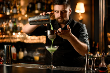 Professional bartender pouring a creamy green color alcoholic drink from the steel shaker to the...