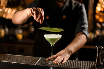 Bartender decorated the green creamy color alcoholic cocktail drink with a green leaf with tweezers