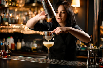 Professional bartender girl pouring a yellow alcoholic drink from the steel shaker to the glass through the strainer filter
