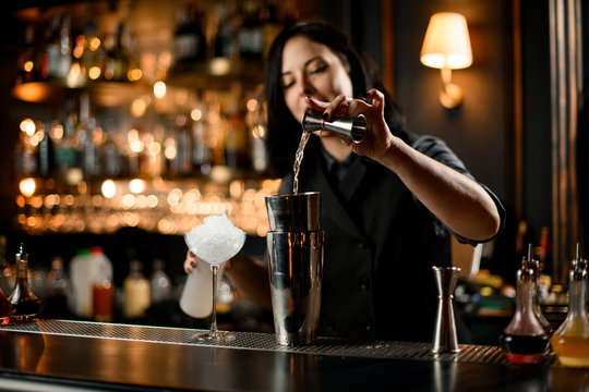 Bartender girl pouring an alcoholic drink from the jigger to a professional steel shaker