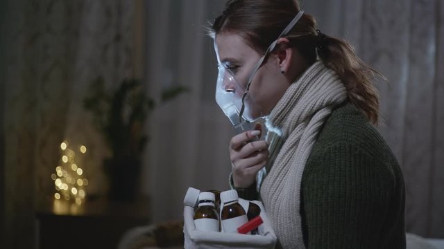 ill woman inhaling through inhaler mask at home, treats inflammation of the airways via nebulizer for preventing asthma and cough sitting on bed with medicines