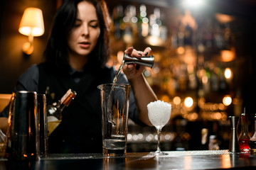 Bartender girl pouring a transparent alcoholic drink from the jigger to a professional steel shaker