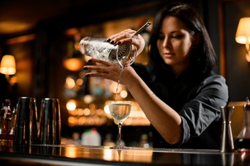 Professional bartender girl pouring a trasparent alcoholic drink from the measuring cup to the...