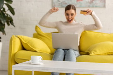 Selective focus of coffee on table and excited woman with credit card and laptop on sofa at background