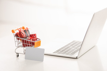 Toy cart with gift boxes beside credit card and laptop on white surface