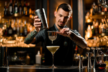 Professional bartender pouring yellow creamy alcoholic drink from the shaker to the glass through the strainer filter