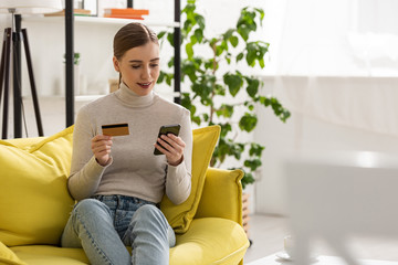 Smiling girl holding credit card and using smartphone on sofa in living room