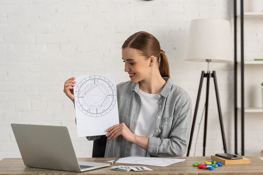 Smiling Astrologer Holding Natal Chart With Cards With Zodiac Signs On Table