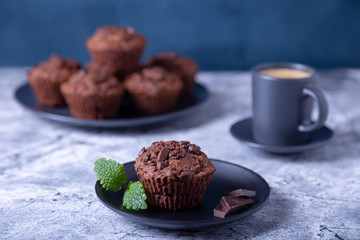 Chocolate muffins with mint on a black plate. Homemade baking. In the background is a cup of coffee and a plate with muffins. Marble table and blue background. 