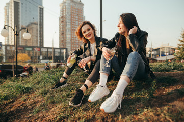 Naklejka na ściany i meble Portrait of happy young girls in leather jackets sitting on grass on cityscape background with bottle of wine and glasses in hands smiling at looking at camera. Girlfriends drink wine on a walk.
