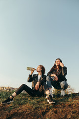 Two stylish girlfriends sitting on a hill on sky background drinking wine at sunset.Two girls on a walk drinking wine and relaxing,sitting on the ground.Fashionable photo of girls drinking wine