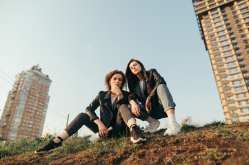 Naklejka na ściany i meble Street photo of stylish girls on sitting on grass on cityscape background with bottle of wine in hands and posing at camera. Two girlfriends in leather jackets on a walk in the city.