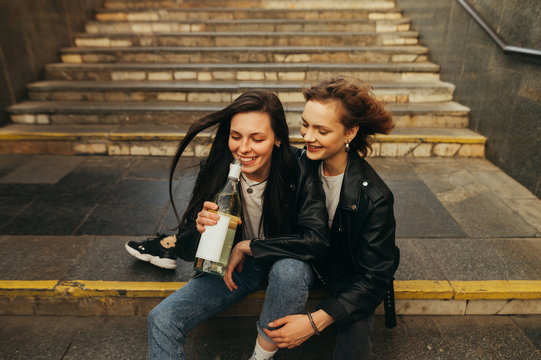 Two happy girls in stylish clothes sit on the stairs in the underground, drinking wine and having fun with smiles. Freedom girlfriends drink wine from a bottle on the street, sitting on the stairs