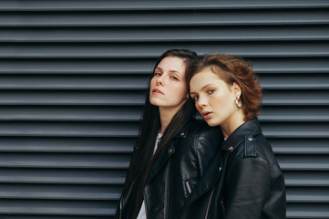 Portrait of two stylish beautiful girls in casual clothes on a black wall background, looking in camera with a serious face. Two models in leather jackets posing for the camera, dark wall background