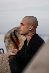 A serious young handsome guy in tattoos on the seashore is standing on the breakwaters. Attractive bald young man with tattoos on his neck by the ocean. Dangerous guy, bully. Stylish man in black.