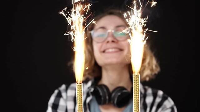 Close up european hipster girl in black and white plaid shirt, headphones on neck celebrating with sparklers and bengal fire. Happy woman smiling girl holds two sparklers close to the camera against