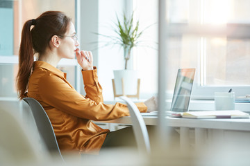 Young woman sitting at her workplace in front of the window with pensive sight and thinking about...