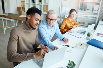 African businessman pointing at laptop and discussing with mature businessman online presentation with young businesswoman working in the background