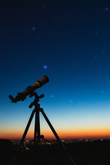 Telescope silhouette and night sky with city lights in the background.