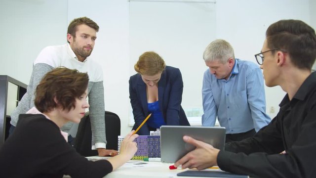 Young man sit at a table with tablet, his workmates look and discuss