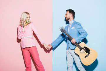 smiling woman with bouquet and handsome man with acoustic guitar holding hands on pink and blue background