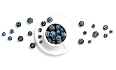 Ripe blueberries in a white cup and a pattern of berries on a white background, top view, flat lay, copy space. Focus on blueberries in a cup.