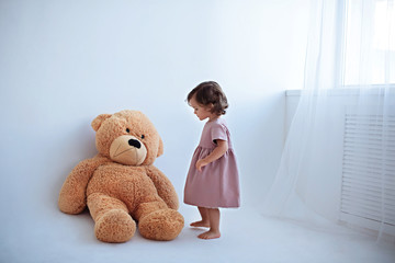 A little girl in a pink dress stands next to a big brown teddy bear in a bright room.