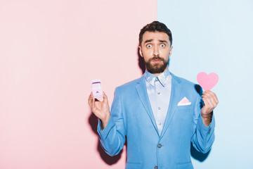 scared man holding heart-shaped card and engagement ring on blue and pink background
