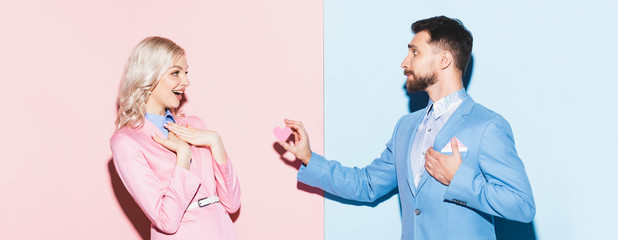 panoramic shot of handsome man giving heart-shaped card to shocked woman on pink and blue background