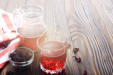 Closeup view at glass of hibiscus ice tea and jug on wooden table background