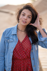 Close up beautiful gorgeous brunette girl in red dress and denim jacket fixing her hair, looking at camera and smiling. Wooden background. Sun set concept.