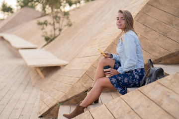 Beautiful smiley blonde girl in denim jacket and skirt sitting on the wood stairs, holding smartphone, looking at camera and the wind developing her hair. Student girl smiling.