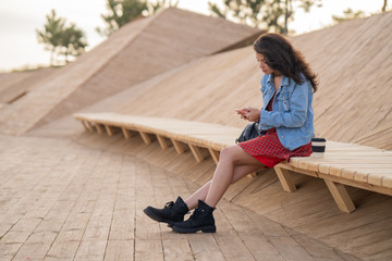 Beautiful gorgeous brunette girl in denim jacket  and red dress  sitting on the wood bench with a eco cup and looking in smartphone.