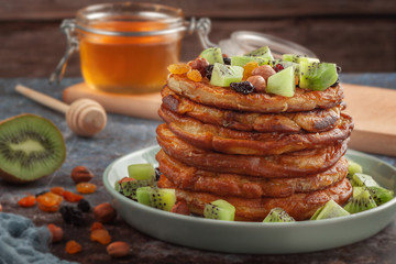 On the table is a dish with pancakes, honey, kiwi, nuts and raisins.