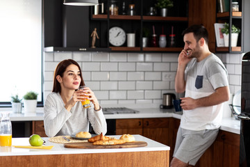 Couple in the kitchen eat breakfast with juice and pastry on table and he is talking on the mobile phone while she drink orange juice