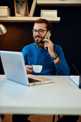 Fototapeta na wymiar Handsome bearded caucasian young entrepreneur sitting in his office, having business call and holding cup of coffee. Start-up business concept.