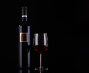 Closed red wine bottle with empty label and glass of wine on black background with reflections. Concept sales, discount price. Photo for advertising