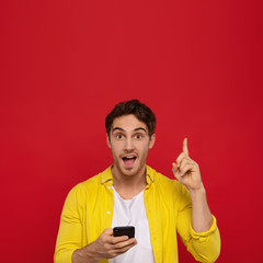 smiling man in yellow shirt using smartphone and pointing with finger up, smiles positively, isolated on red background. Good mood concept