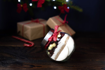 Biscuit ingredients: flour, sugar, dried cranberries, white chocolate, are laid out in layers in a jar, with a red ribbon standing on a wooden table. In the background a fir branch with gifts.