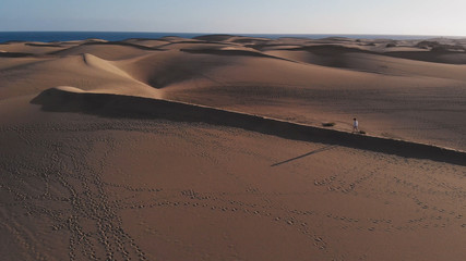 Breathtaking view from a height - a beautiful girl walks through the desert, on the ridge of the dunes in a strong wind, AERIAL. Maspalomas, Gran Canaria