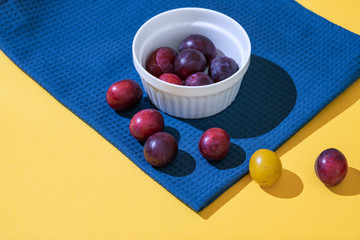 A many ripe dark blue plum stands in a white plate on a blue fabric. Harvest lies on the table. Fruit minimal concept on a yellow background