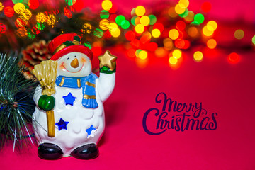 Holiday greeting card. Toy Christmas snowman with star on a red background. Composition of Christmas tree, berries, Apple and colorful bokeh lights garland. Classic atmosphere of new year. Copy space